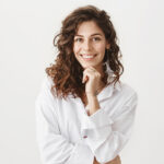 cute happy european woman with curly hair in trendy white blouse, smiling broadly, leaning chin on hand, listening with interest to friend story, standing amused and joyful over gray wall